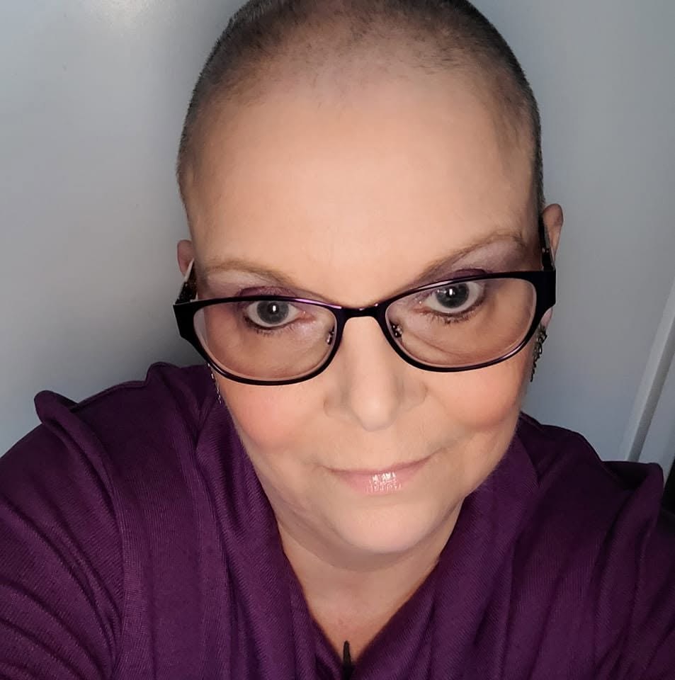 Debbie smiling shortly after shaving her head, wearing purple clothing and makeup.